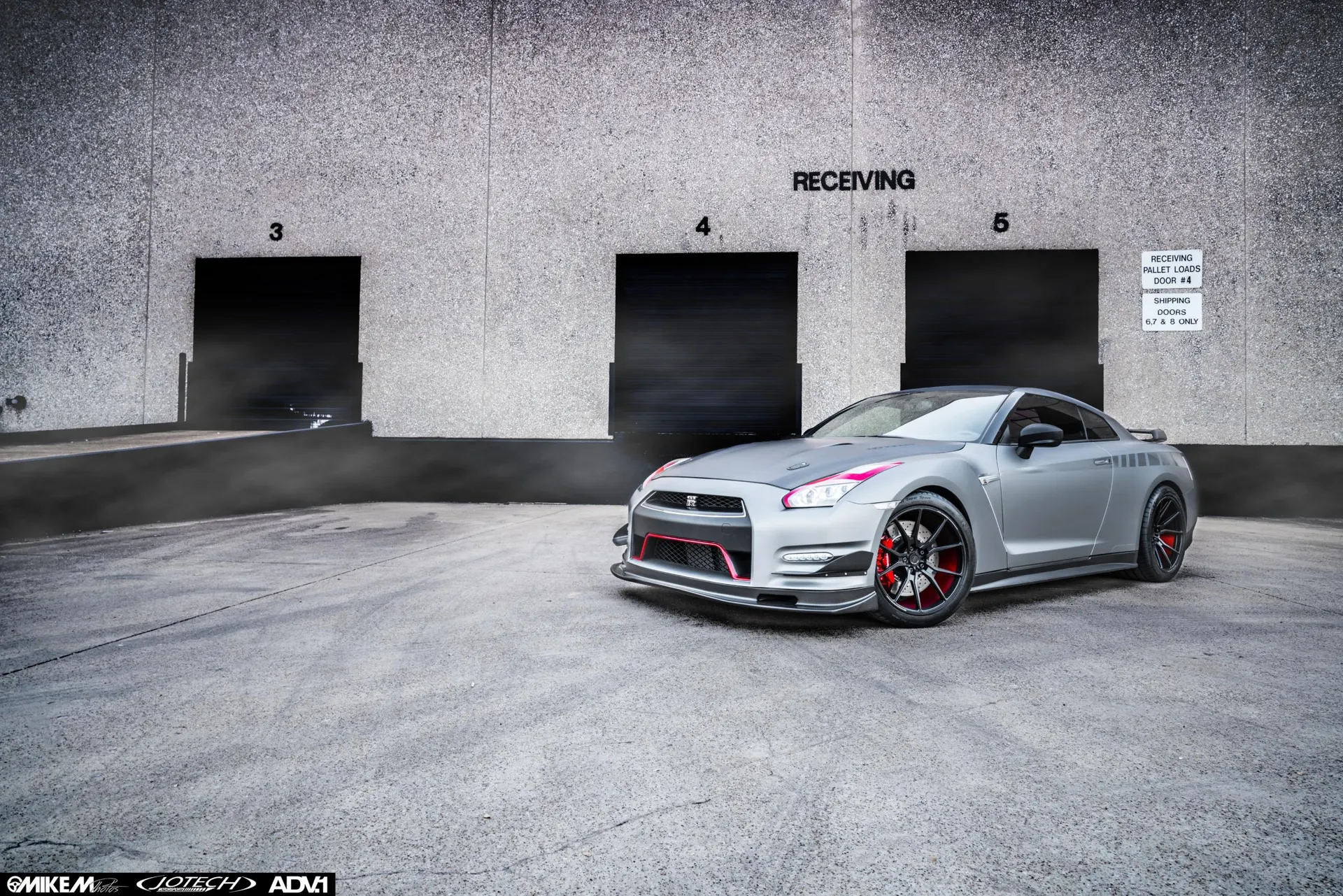 A silver Nissan GT-R with red accents and ADV.1 wheels parked in front of a warehouse loading dock.