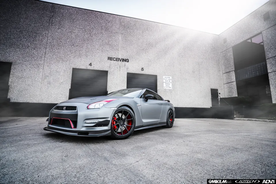 A matte grey Nissan GT-R with red accents and aftermarket wheels parked in front of a concrete industrial building.