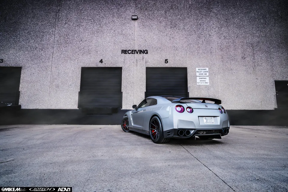 Silver Nissan GT-R R35 with aftermarket wheels and a large rear wing parked in front of a warehouse receiving dock.