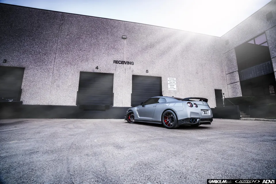 Grey Nissan GT-R with aftermarket wheels and exhaust parked in front of a receiving dock at an industrial building.