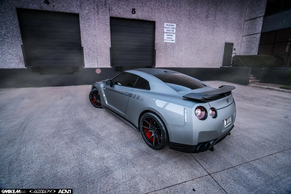 Grey Nissan GT-R with aftermarket wheels, red brake calipers, and a rear wing, parked at a loading dock.