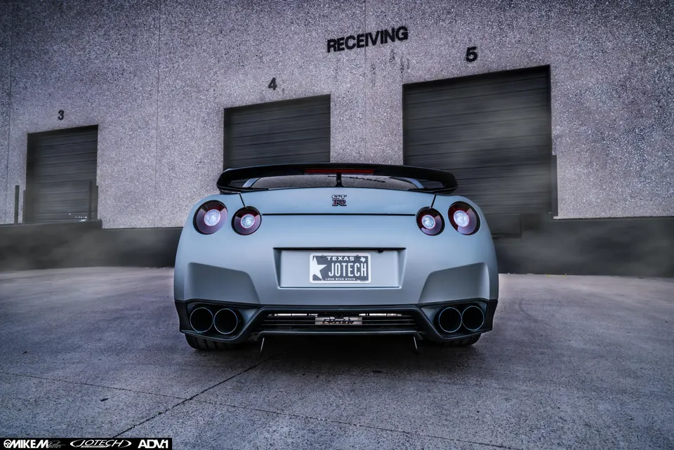 Rear view of a grey Nissan GT-R R35 with quad exhaust tips and a rear wing, parked in front of a warehouse.