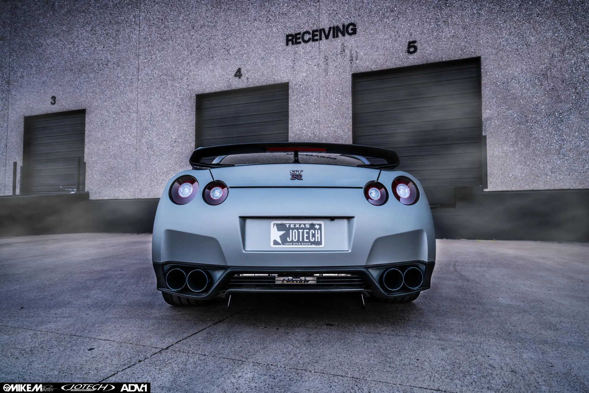 Rear view of a grey Nissan GT-R R35 with quad exhaust tips and a rear wing, parked in front of a warehouse.