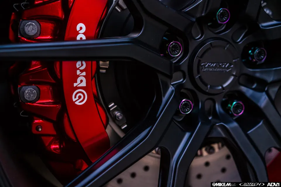 Close-up of a black multi-spoke wheel with iridescent lug nuts and a red Brembo brake caliper and drilled rotor.