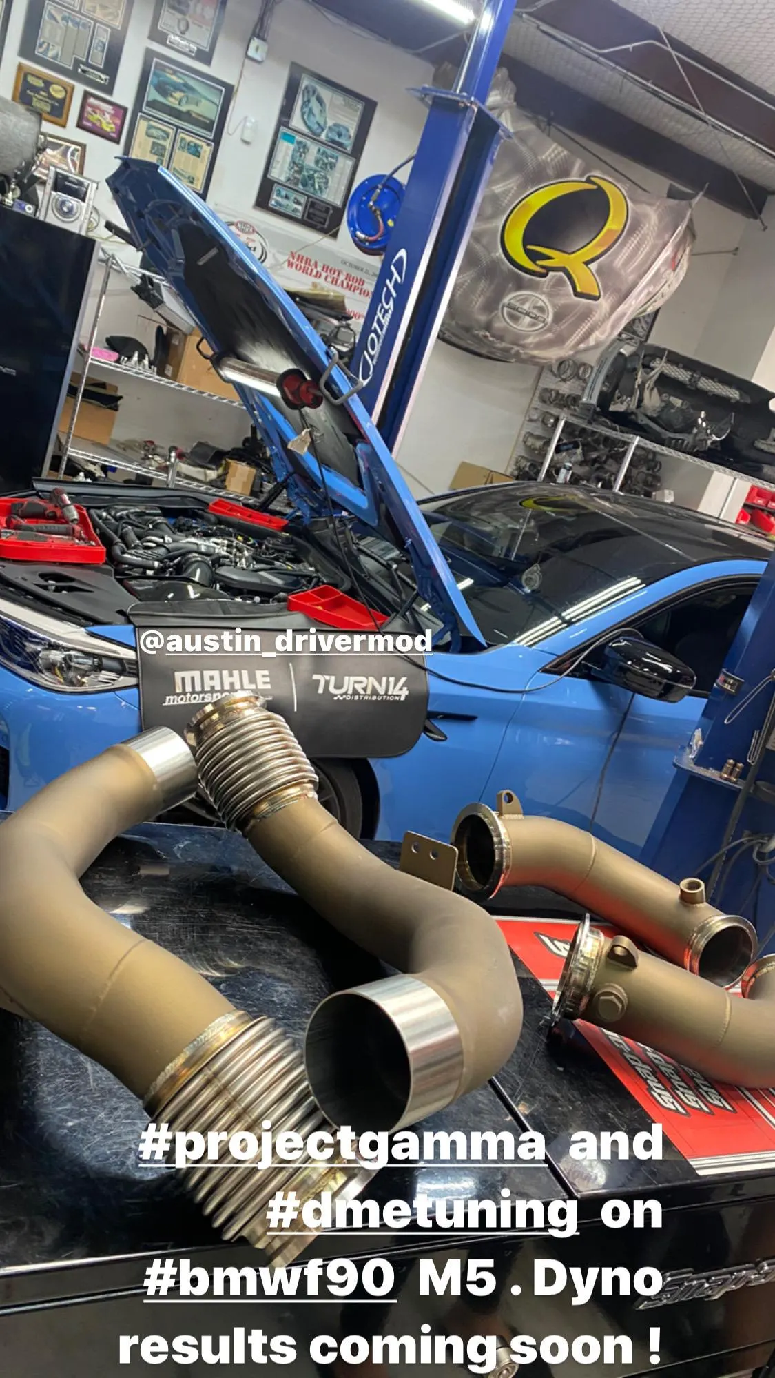 A blue BMW F90 M5 with its hood open on a lift in a performance shop, with aftermarket exhaust downpipes on a workbench.