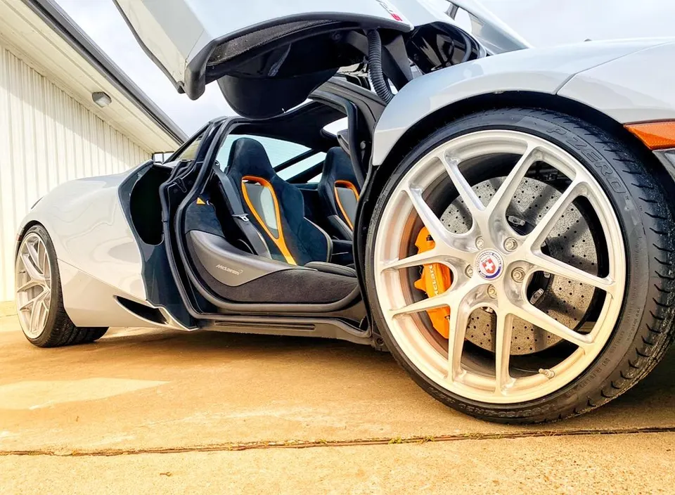 A McLaren supercar with butterfly doors open, showcasing custom silver HRE wheels, orange brake calipers, and a black and orange interior.
