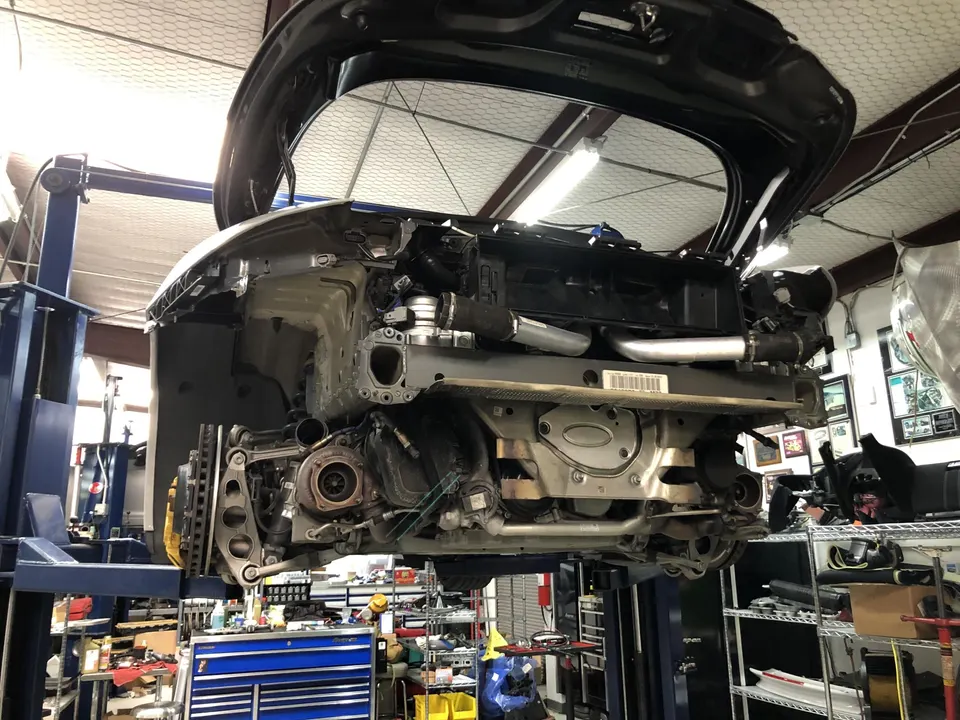 Rear view of a Porsche 911 on a lift in a performance shop, with the bumper removed exposing twin turbochargers and exhaust.