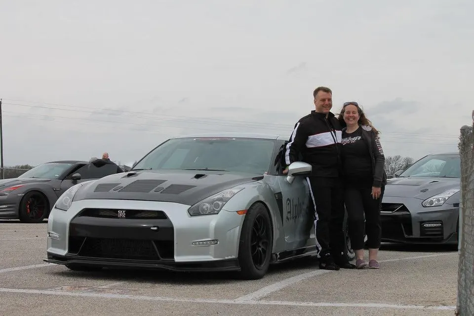 A smiling couple stands next to a silver Nissan GT-R with a black hood at a track event, with other GT-Rs in the background.