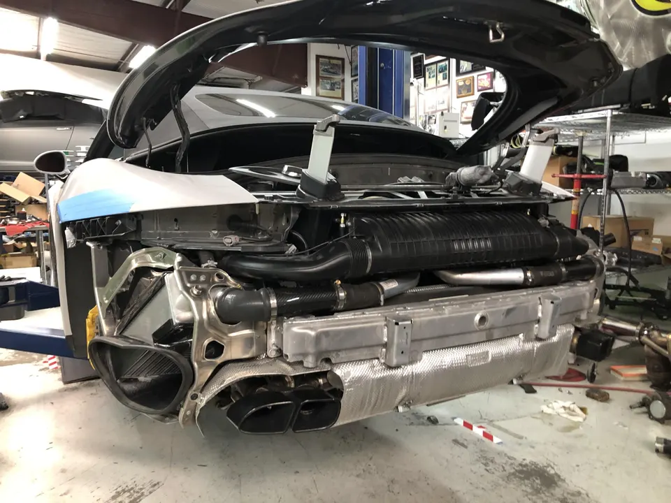 Porsche 911 in a shop with rear bumper removed, showing exhaust system, intercooler, and airbox during performance upgrade.