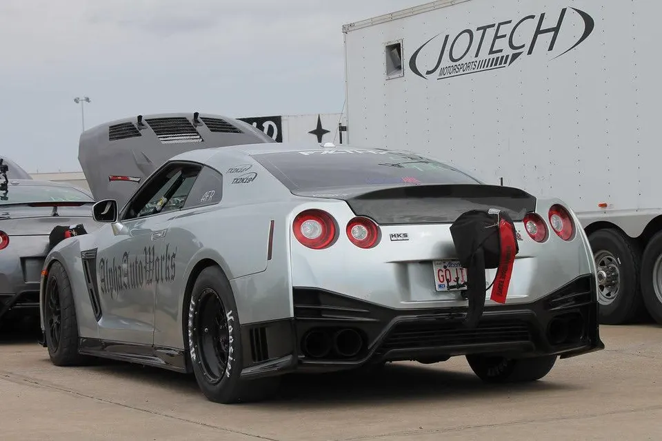 Silver Nissan GT-R drag car with a parachute and Hoosier drag tires parked at a track, next to a Jotech Motorsports trailer.