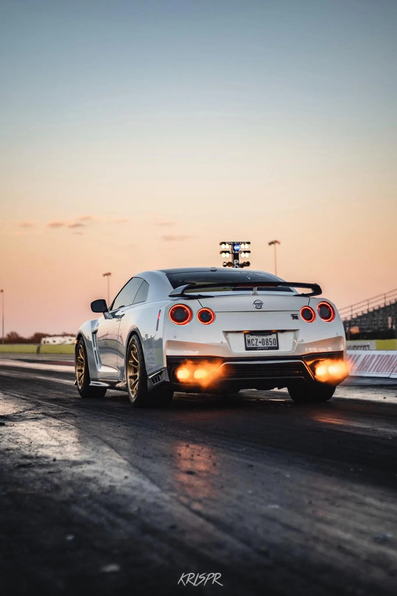 A white Nissan GT-R with flames shooting from its exhaust on a drag strip at sunset, ready to launch.
