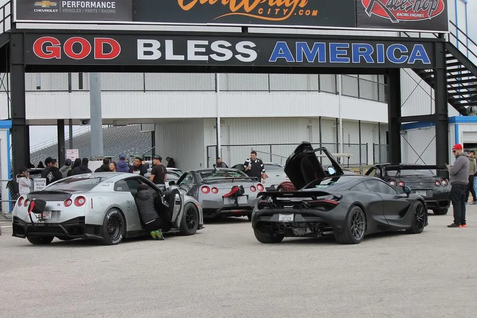 Nissan GT-Rs and a McLaren 720S in the staging lanes at a drag strip event under a 'God Bless America' sign.