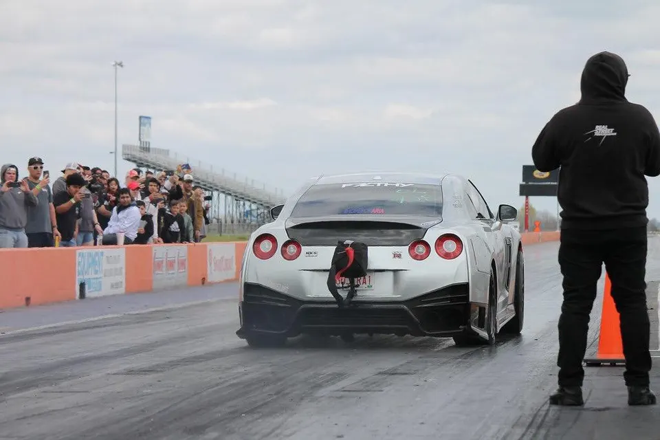 A silver Nissan GT-R with a parachute attached to the rear, staged at a drag strip with spectators watching.