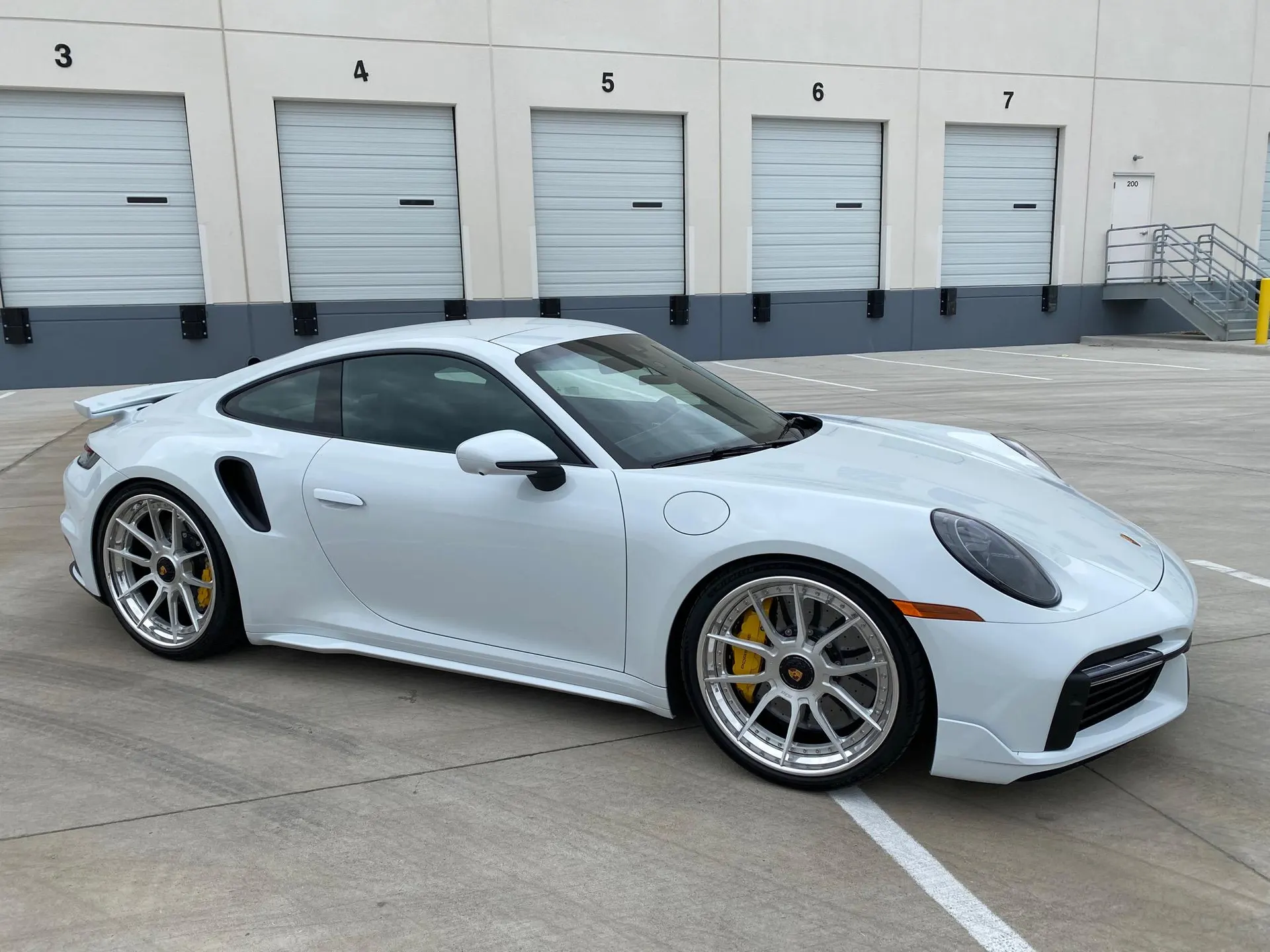 A white Porsche 911 Turbo S with custom silver wheels and yellow brake calipers parked in an industrial lot.