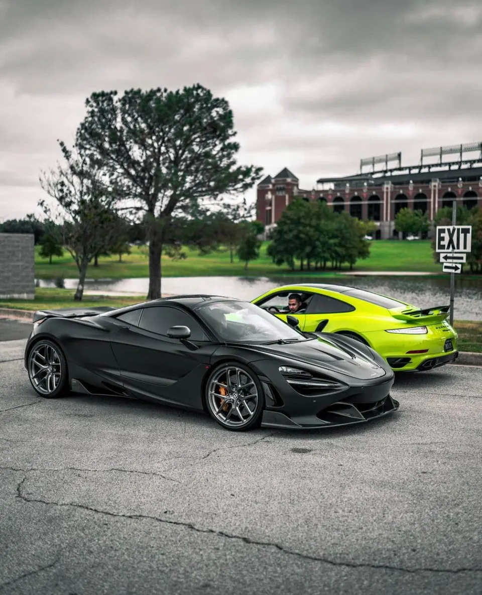 Black McLaren 720S and lime green Porsche 911 GT3 RS parked outdoors with a building and lake in the background.