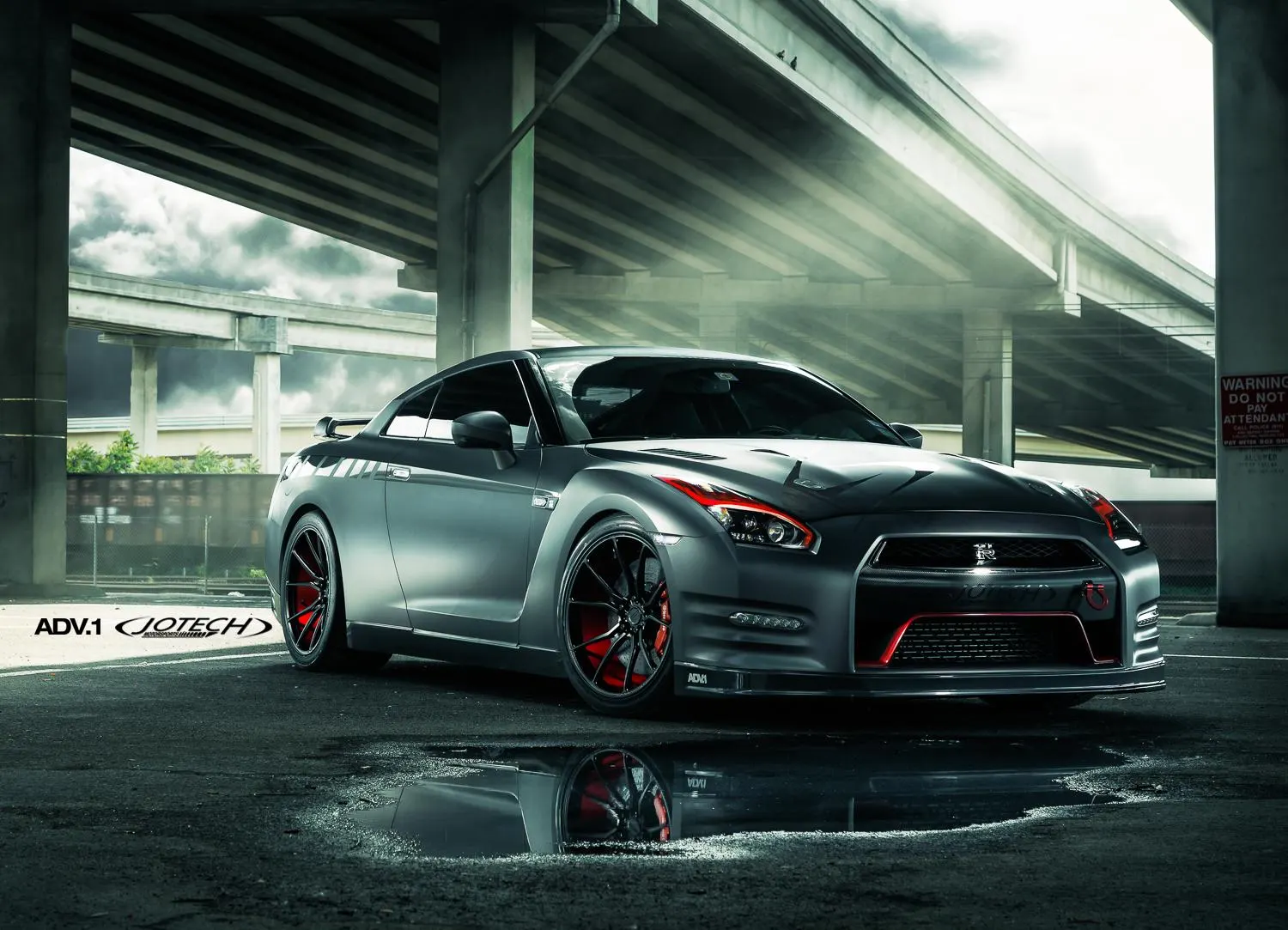 A modified Nissan GT-R with ADV.1 wheels and red accents parked under an overpass, reflecting in a puddle.