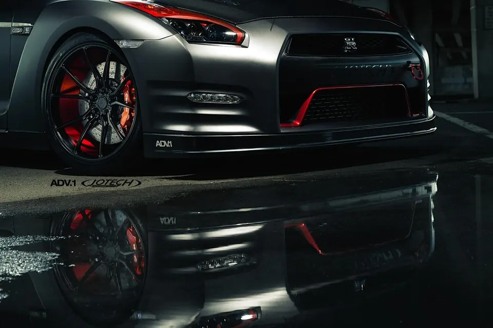A matte black Nissan GT-R with ADV.1 wheels and red brake calipers reflected in a puddle.