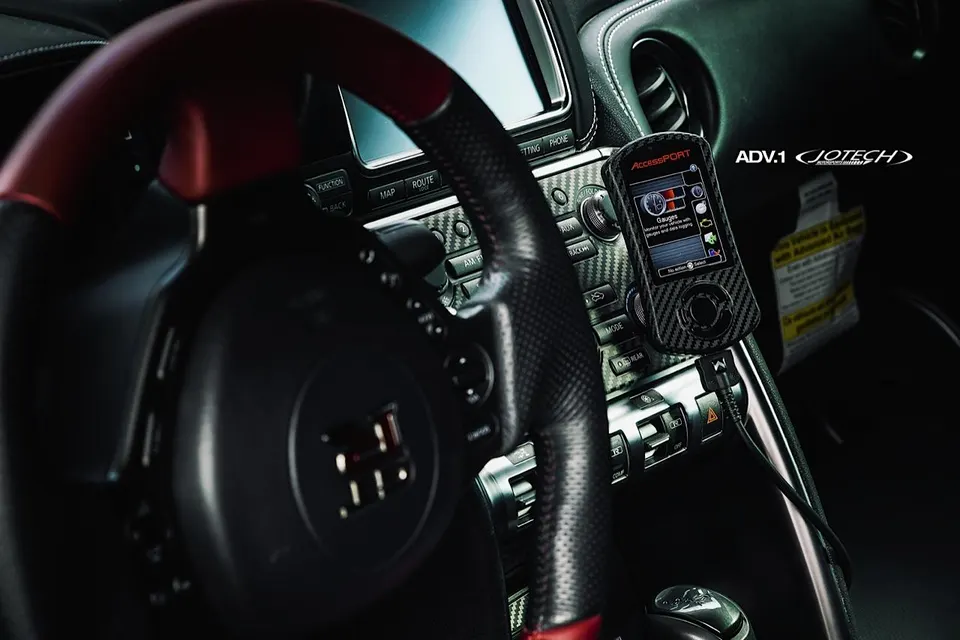 A Cobb AccessPORT tuning device mounted on the carbon fiber dashboard of a Nissan GT-R interior.