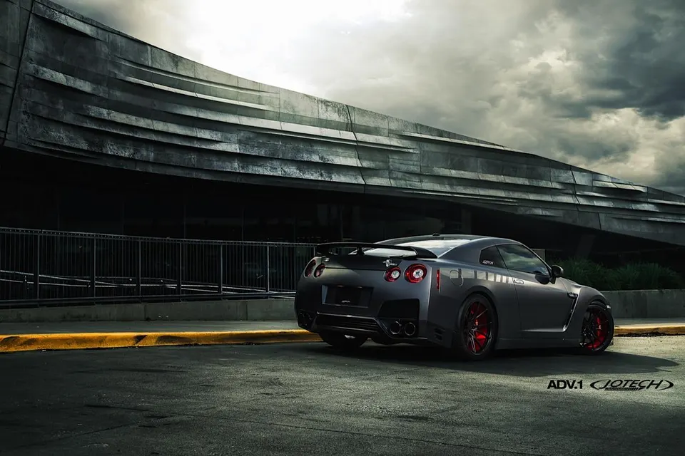 A grey Nissan GT-R with red ADV.1 wheels parked in front of a modern building under a cloudy sky.
