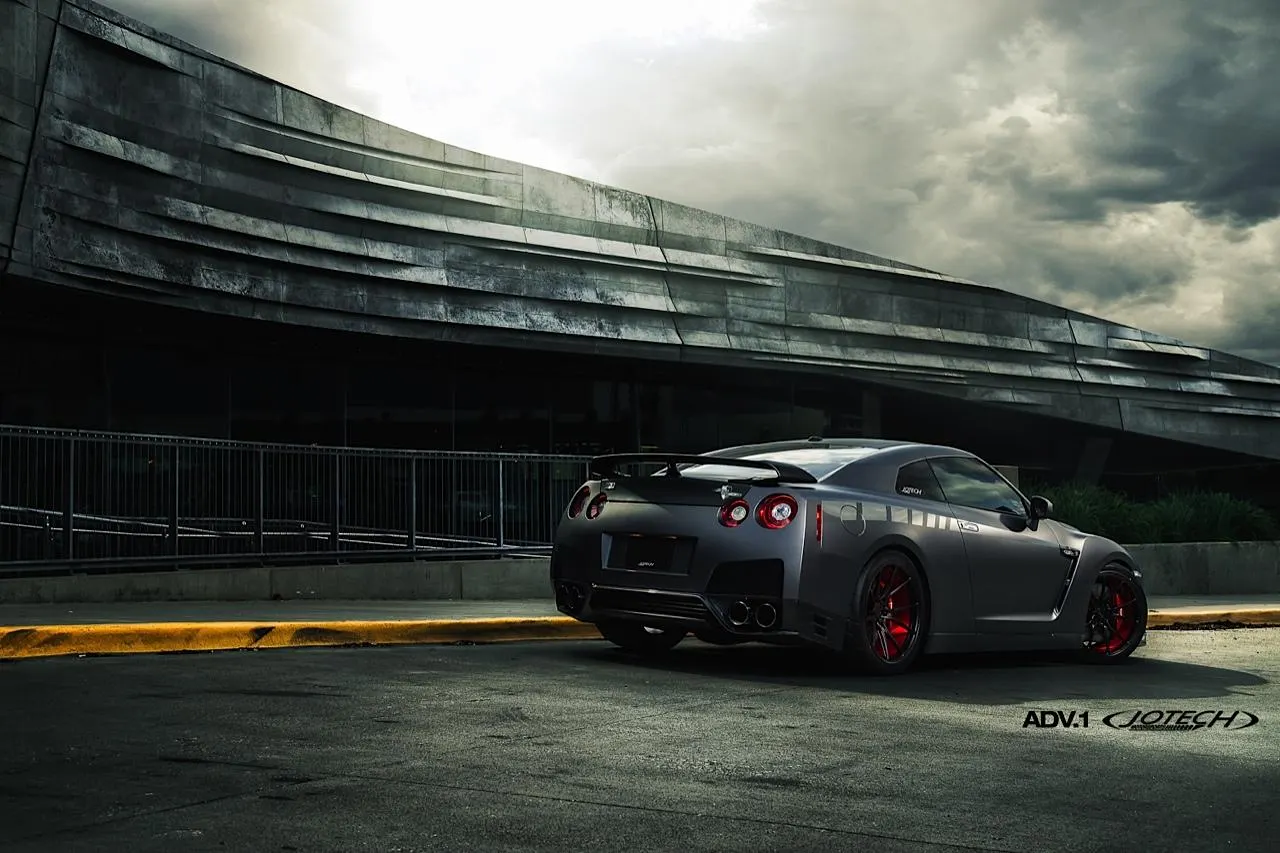 A grey Nissan GT-R with red ADV.1 wheels parked in front of a modern building under a cloudy sky.