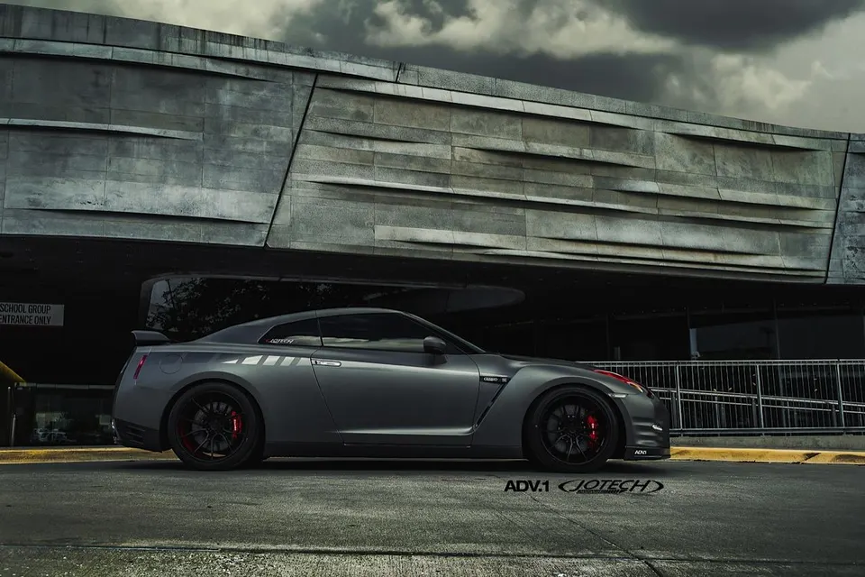 Grey Nissan GT-R with ADV.1 wheels parked in front of a modern concrete building under a cloudy sky.