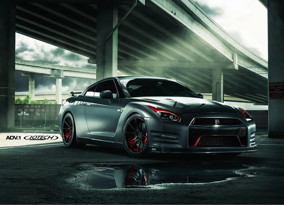 A custom gray Nissan GT-R with red accents and ADV.1 wheels parked under an overpass, reflecting in a puddle.