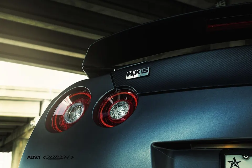 Close-up of the rear of a matte black Nissan GT-R, showcasing its round taillights, carbon fiber trunk trim, and HKS badge.