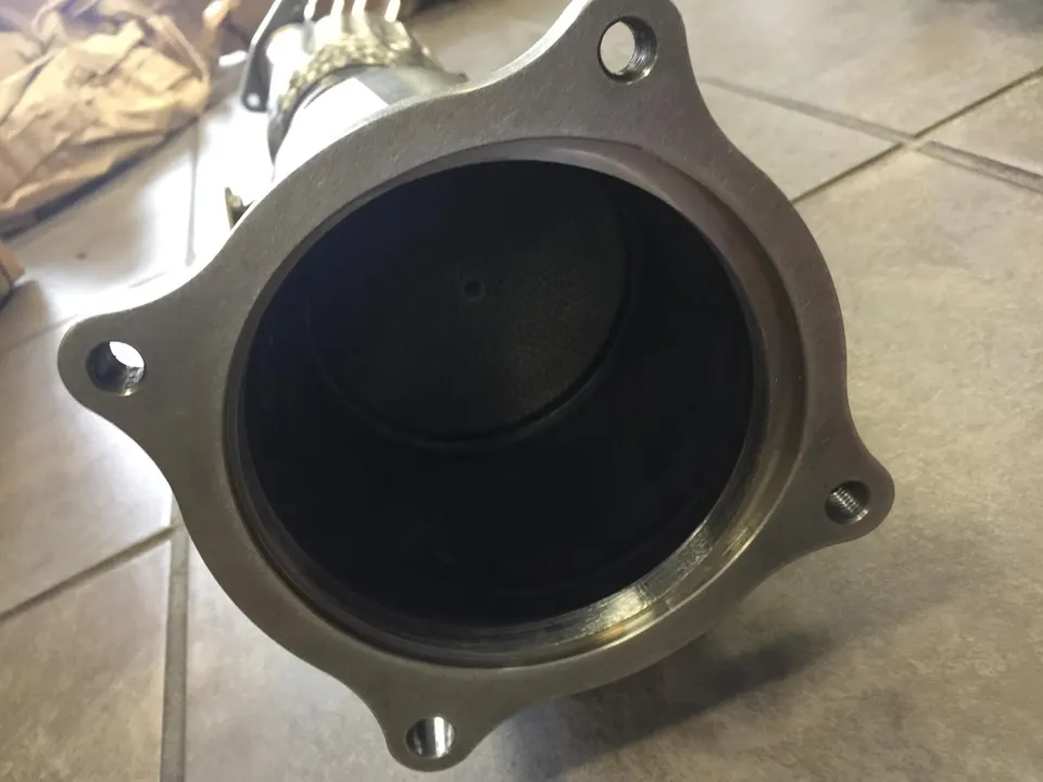 Close-up view of a new performance exhaust downpipe flange, showcasing the metal construction and bolt holes on a tiled floor.