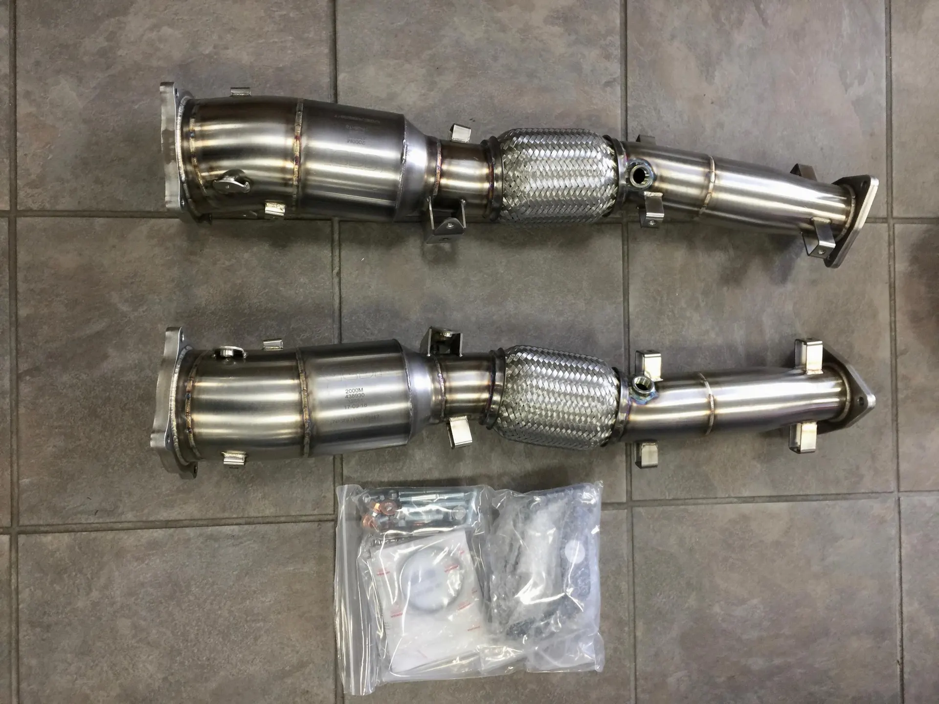 Two new stainless steel aftermarket exhaust downpipes with flexible sections and installation hardware on a tiled floor.