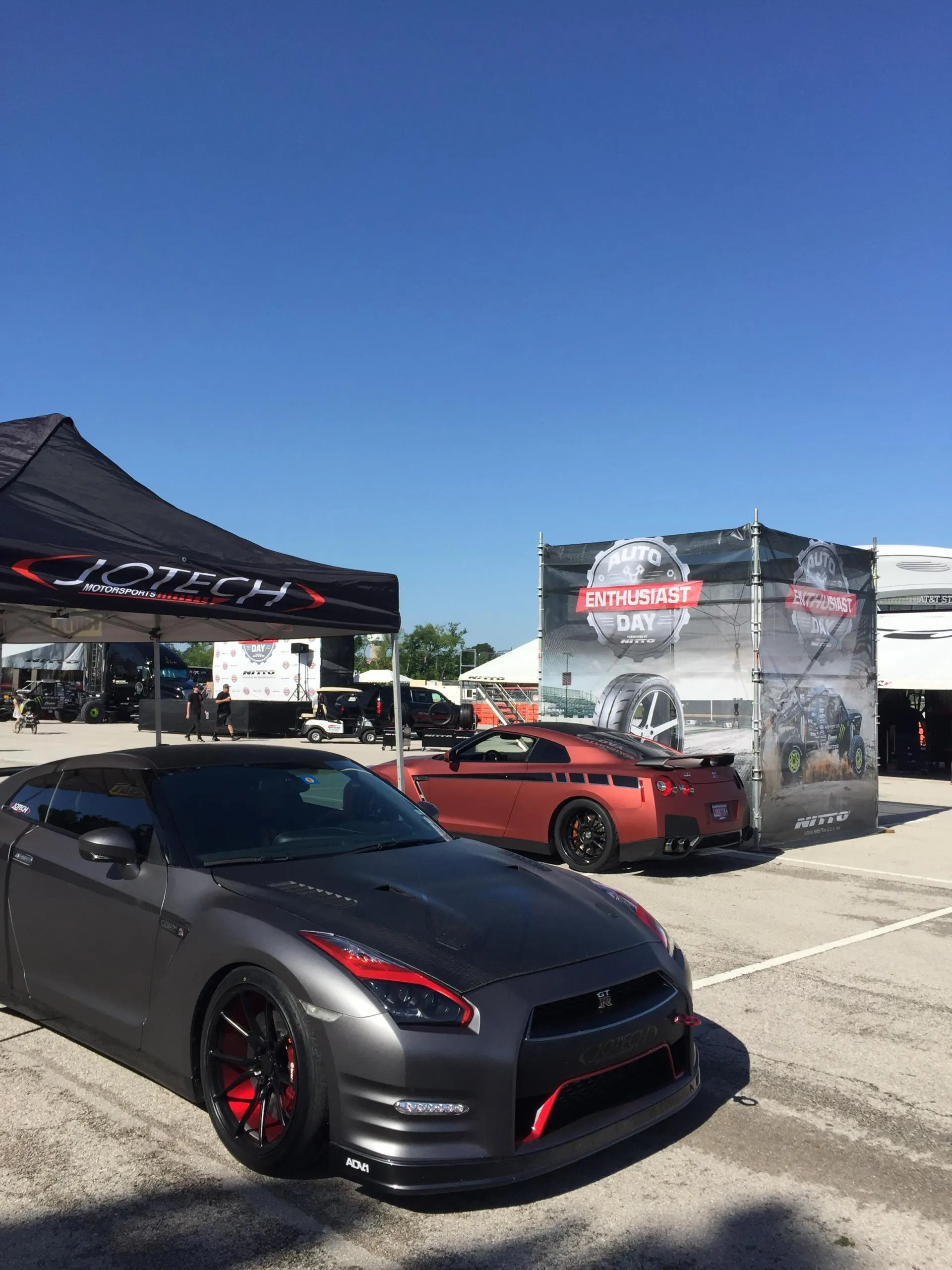 Two modified Nissan GT-Rs, one gray and one red, parked at an outdoor car enthusiast event with Jotech Motorsports tent.
