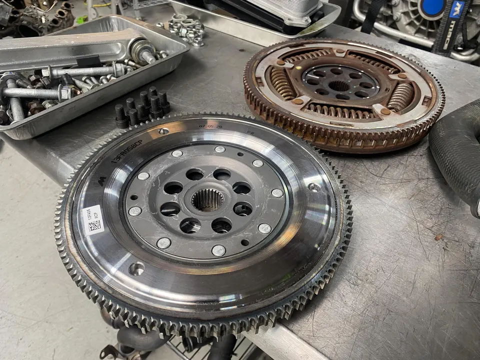 New and old flywheels on a metal workbench in a performance tuning shop, alongside a tray of bolts.