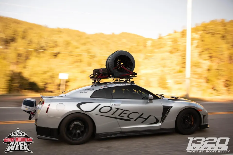 Silver Jotech Nissan GT-R with a roof rack and parachute driving on a scenic road during Rocky Mountain Race Week.