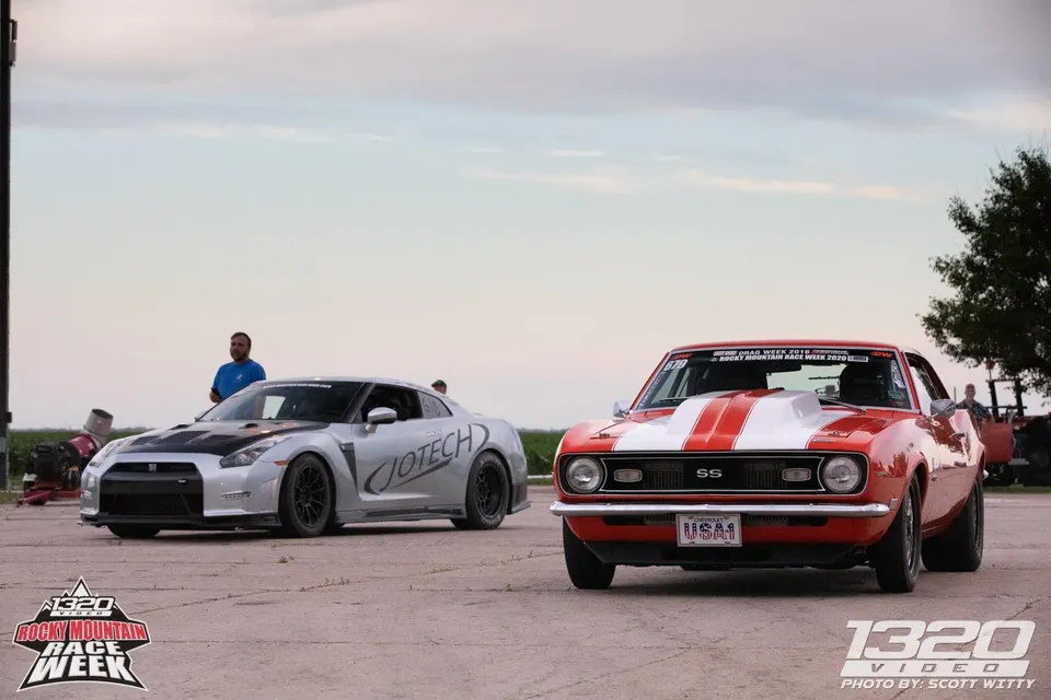 A silver Nissan GT-R with Jotech branding and a red and white classic Chevrolet Camaro at a drag racing event.