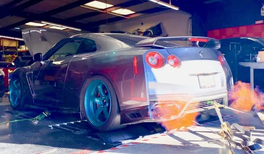 A grey Nissan GT-R on a dyno with bright orange flames shooting from its exhaust during a high-performance test.