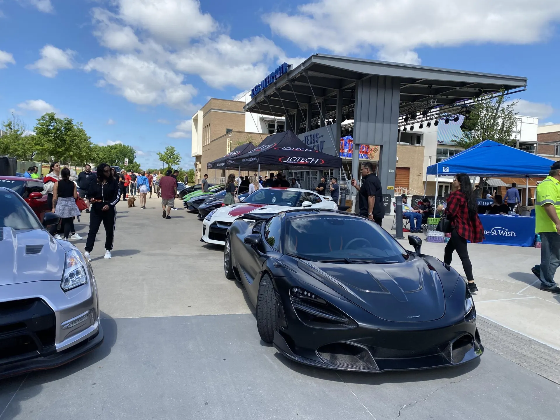 A black McLaren 720S and a silver Nissan GT-R parked at an outdoor car show with many people and tents.