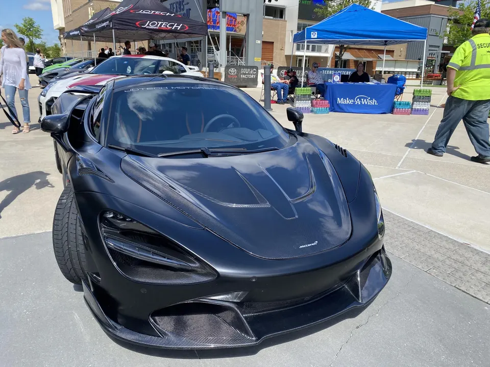 A black McLaren 720S with carbon fiber body parts parked at an outdoor car show event with people and tents in the background.