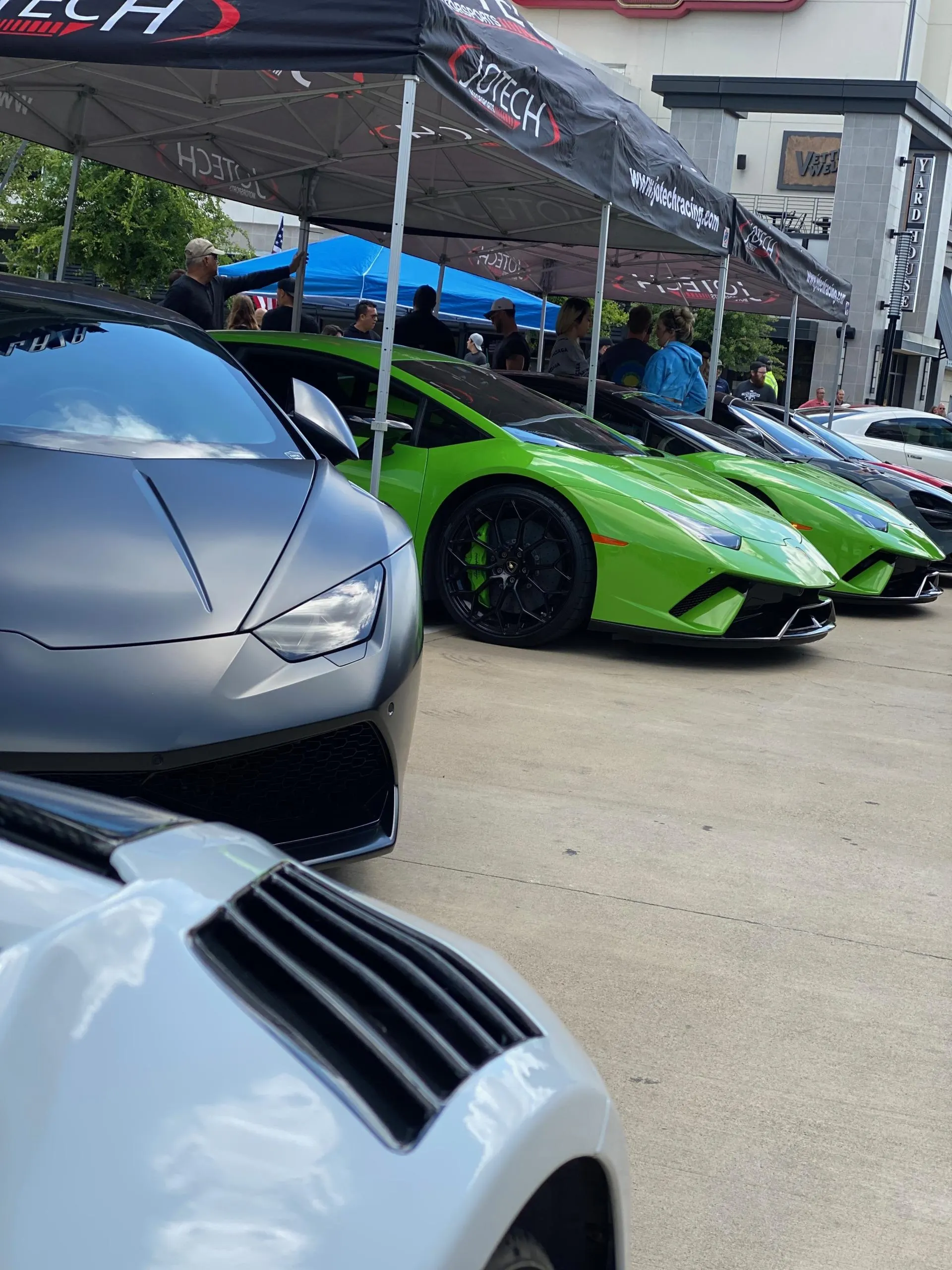 Multiple high-performance Lamborghinis, including green and grey models, parked under Jotech Motorsports tents at a car event.