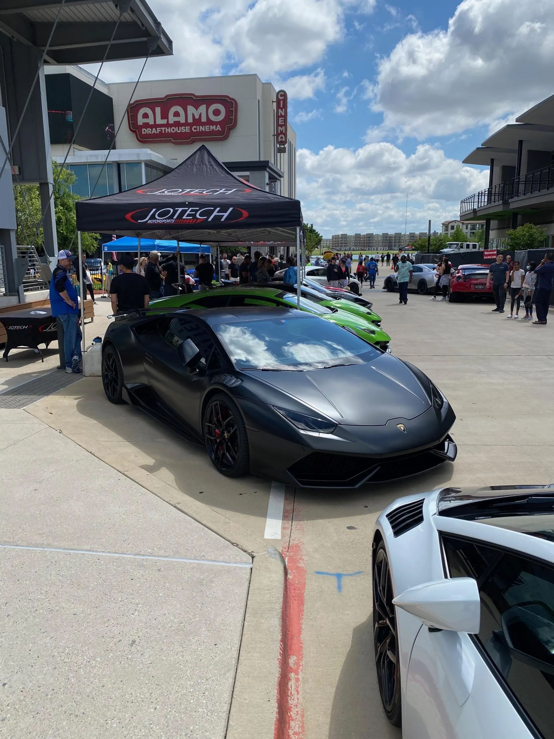 A dark grey Lamborghini Huracan parked at a Jotech Motorsports car meet in front of Alamo Drafthouse Cinema, with a crowd of people.