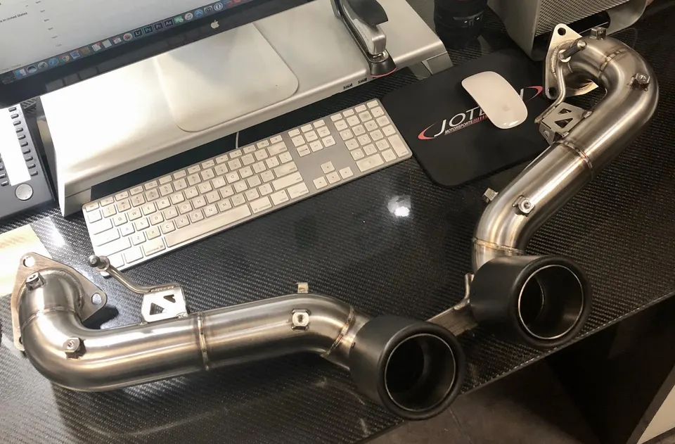 Aftermarket stainless steel exhaust system with black tips displayed on a carbon fiber desk next to a computer.