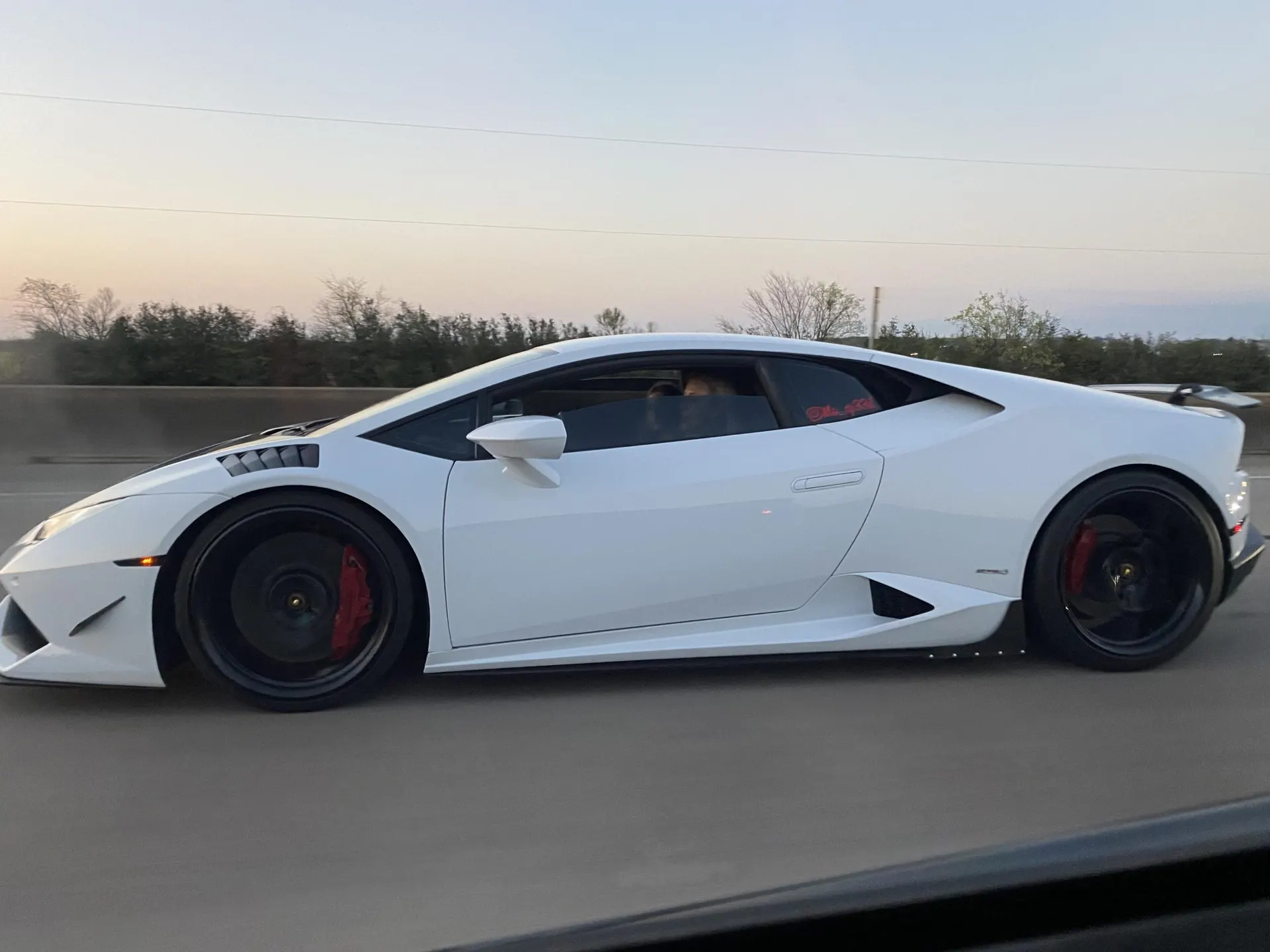 A white Lamborghini Huracan with an aftermarket aero kit and black wheels driving on a highway at sunset.