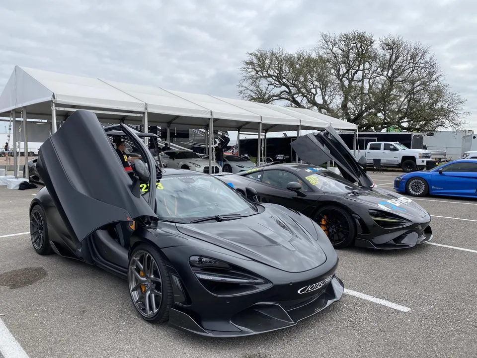 Two black McLaren 720S supercars with open doors parked under tents at a track event paddock.