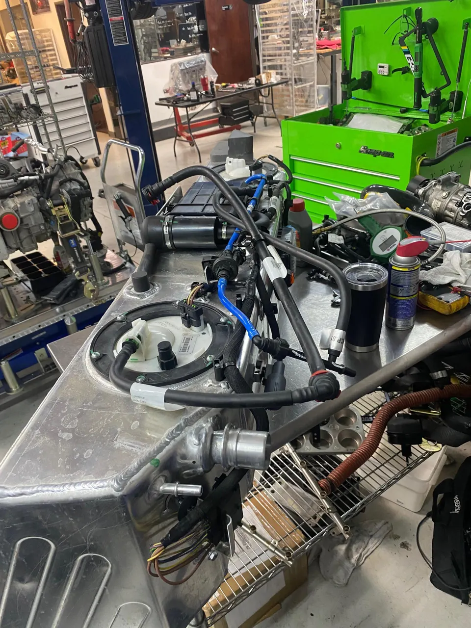 A custom aluminum fuel cell with fuel pump assembly and lines on a workbench in a performance tuning shop.