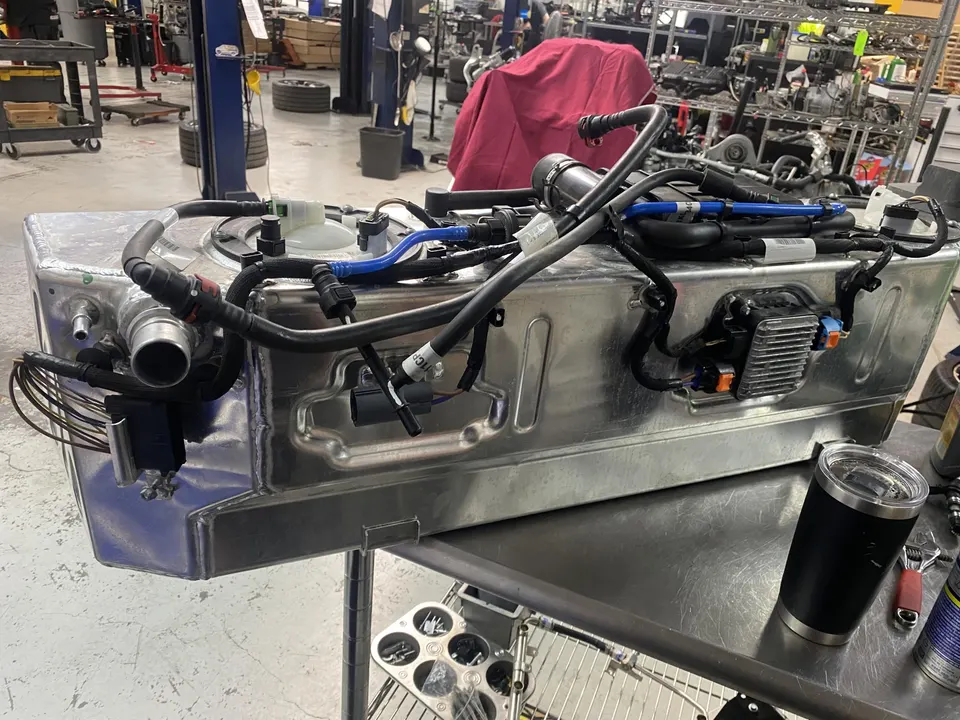Aftermarket fuel tank with fuel lines and electronics on a workbench in a performance tuning shop.