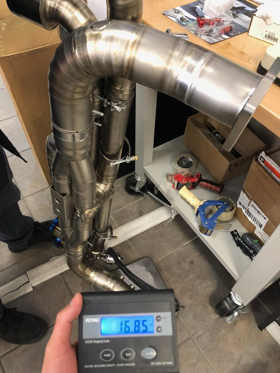A person weighing a large, multi-pipe titanium exhaust system on a digital scale in a workshop.