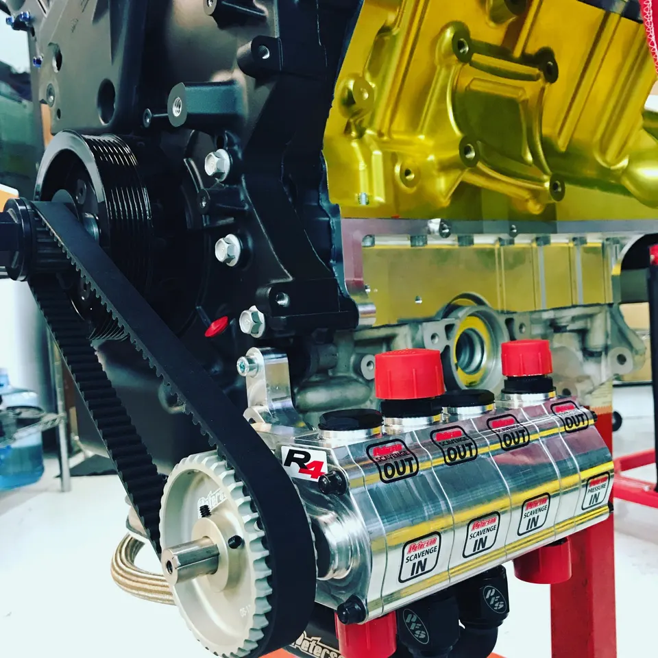 Close-up of a high-performance engine build featuring a black engine block, gold valve cover, timing belt, and Peterson R4 dry sump system.