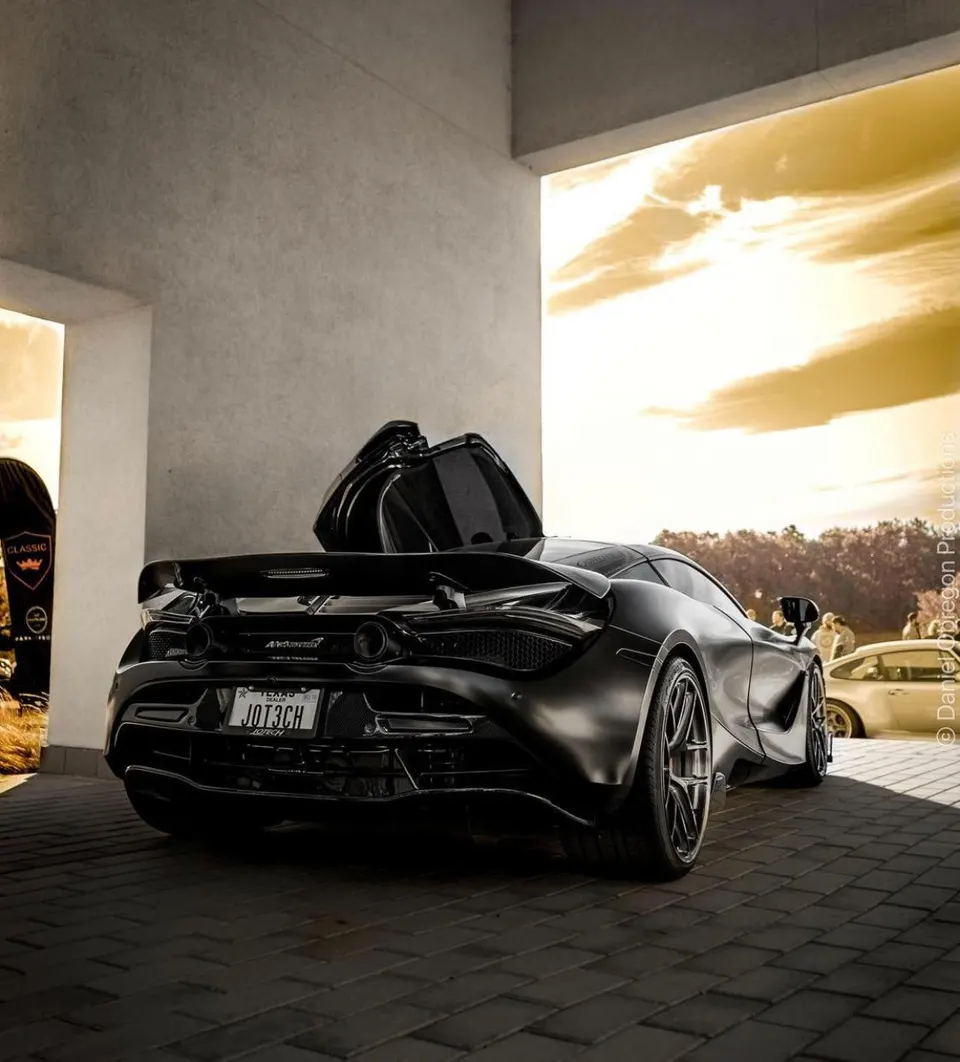 Rear view of a black McLaren 720S with its active aero wing deployed, parked on a paved surface under a dramatic sky.