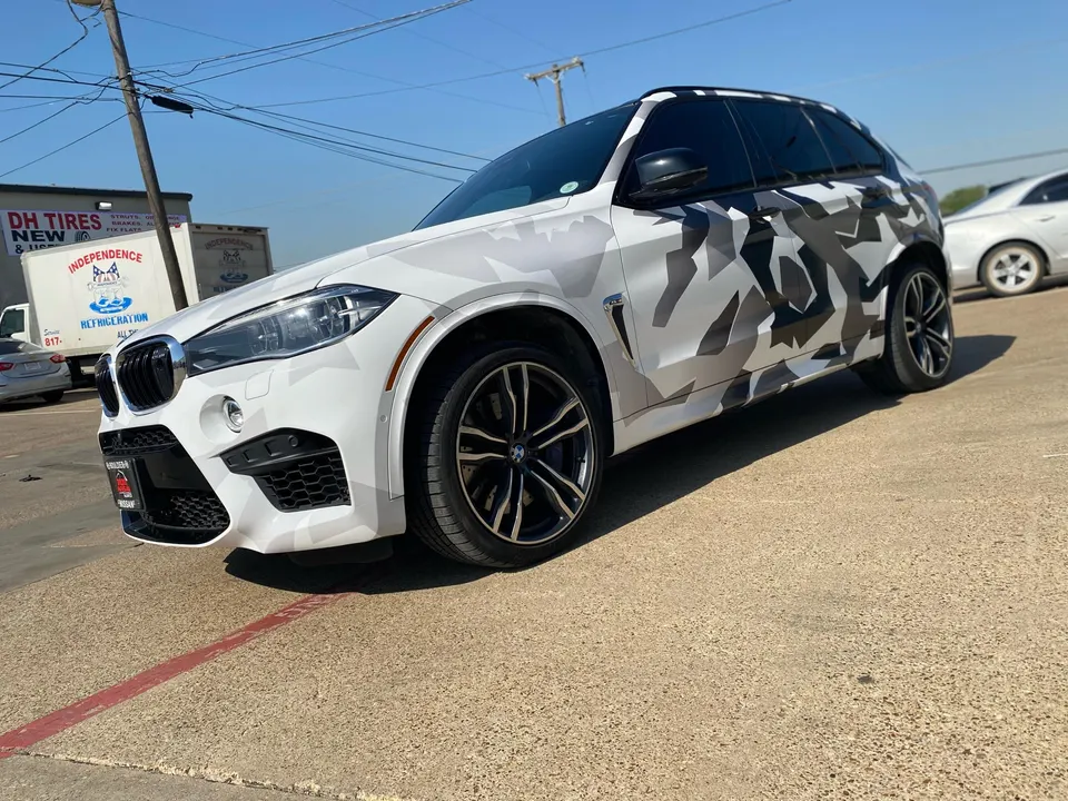 A BMW X5 SUV with a white and black camouflage wrap parked outdoors on a sunny day.