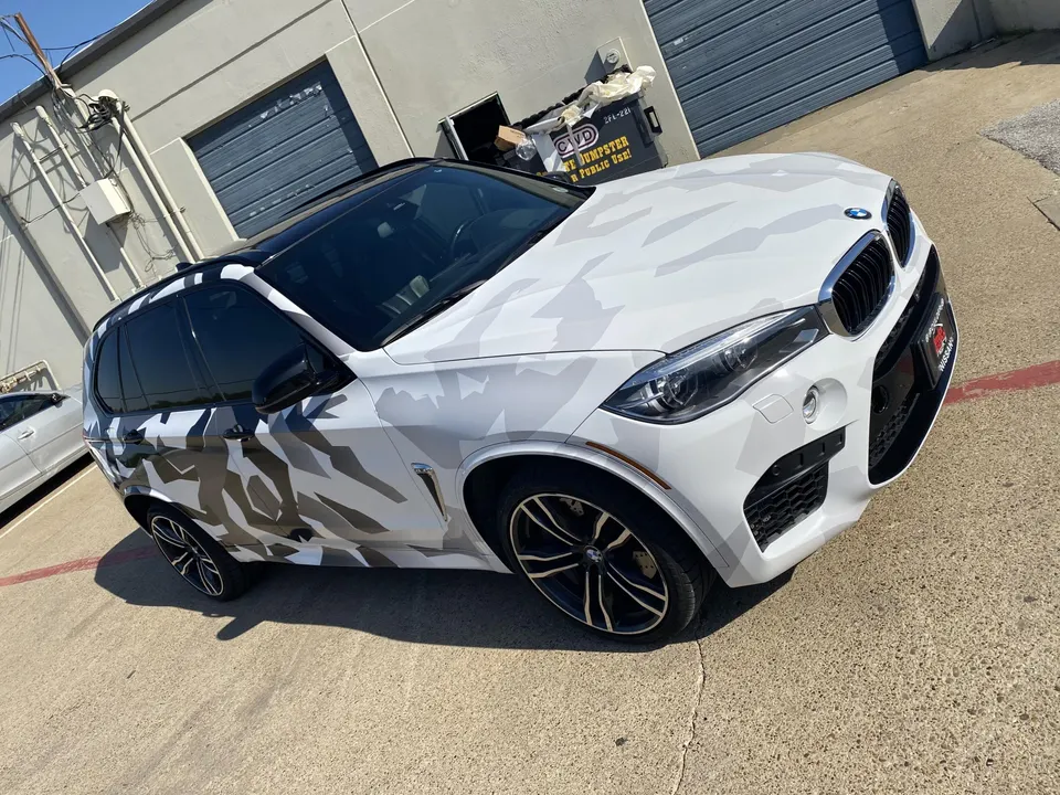 A BMW X5 SUV with a white and grey camouflage wrap parked outside a building on a sunny day.