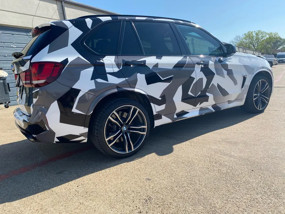 A BMW X5 SUV with a black, white, and grey camouflage wrap parked outdoors on a sunny day.