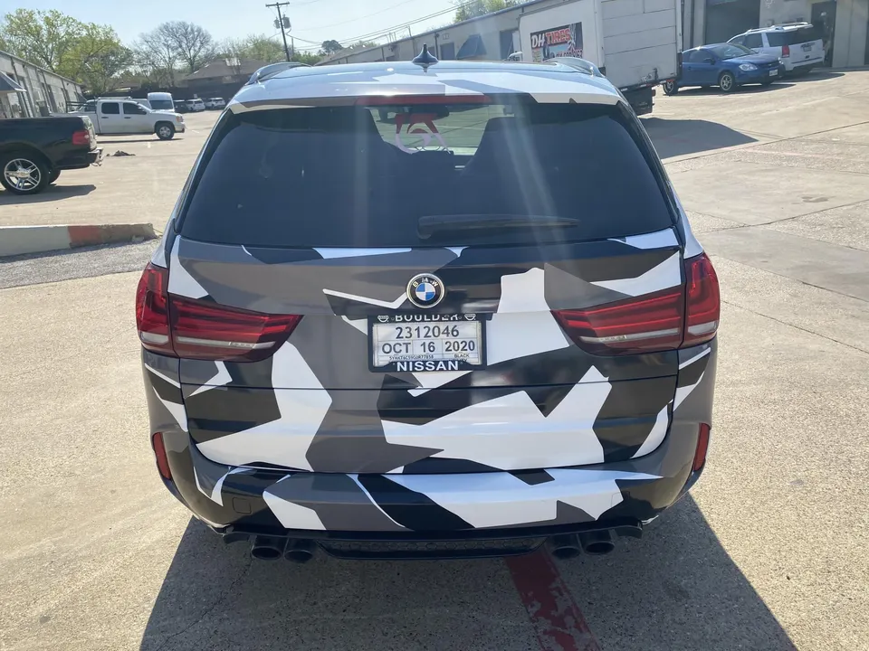 A BMW X5 SUV with a black, white, and grey camouflage wrap, featuring custom exhaust tips, parked outdoors.
